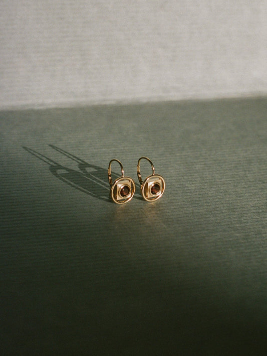 Gold earrings by Rebecca Mapes with a central gemstone on a dark surface