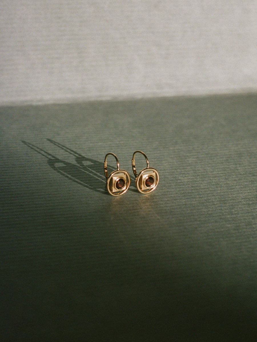 Gold earrings by Rebecca Mapes with a central gemstone on a dark surface